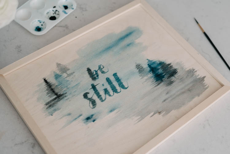 Be Still hand lettering over foggy trees watercolour painting on wood