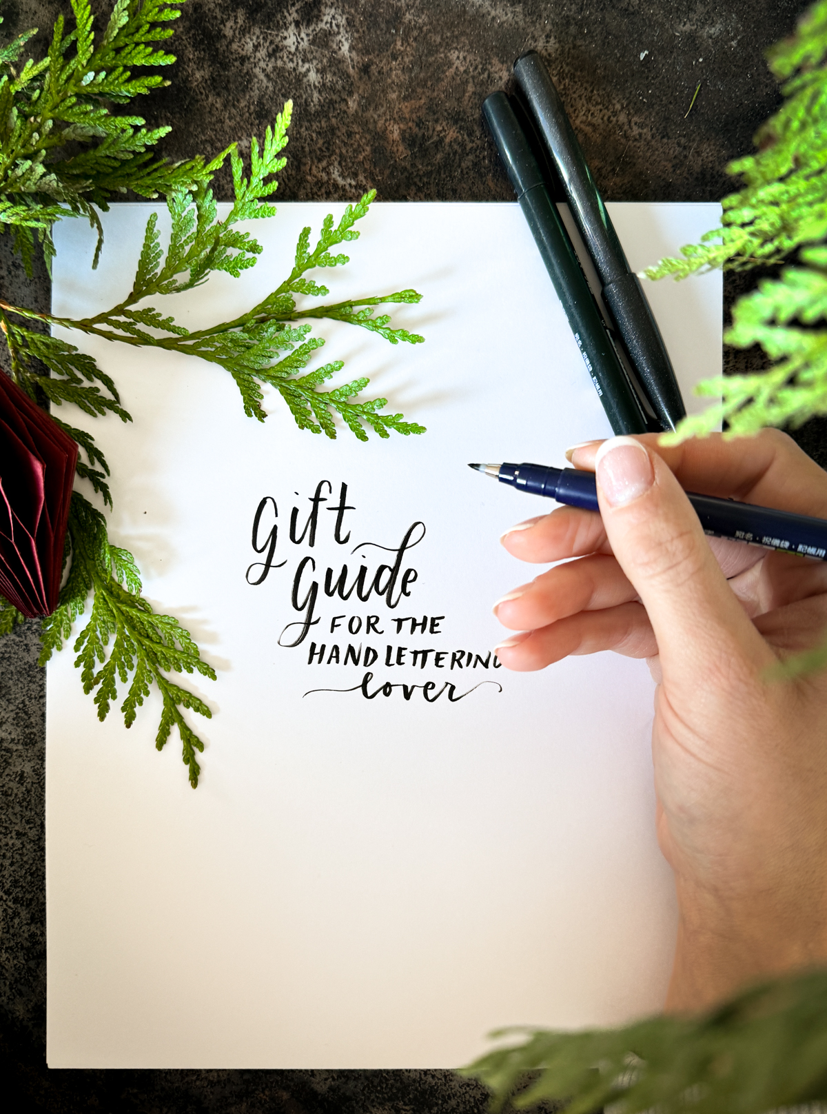 hand lettered words on paper surrounded by cedar branches and a holiday ornament. lettering on paper reads: gift guide for the hand lettering lover. Hand holding small brush pen in photo