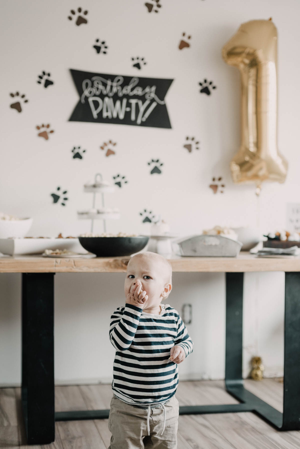 First birthday party with a puppy theme- paw prints backdrop and puppy balloon, 1 year old standing in front of party table