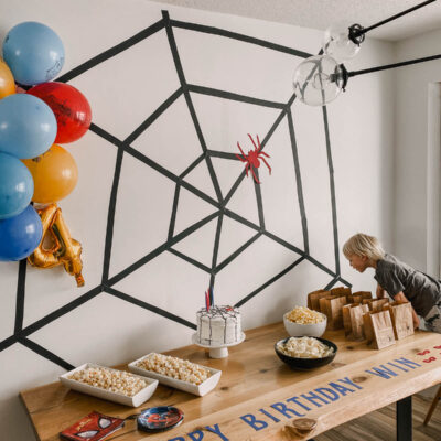 photo of a spiderman themed birthday party table with spiderman balloons, a giant spiderweb backdrop and a table full of party snacks and cake