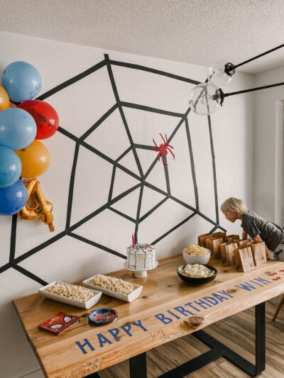 photo of a spiderman themed birthday party table with spiderman balloons, a giant spiderweb backdrop and a table full of party snacks and cake