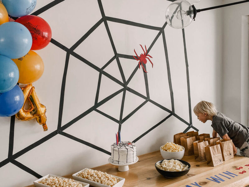 photo of a spiderman themed birthday party table with spiderman balloons, a giant spiderweb backdrop and a table full of party snacks and cake