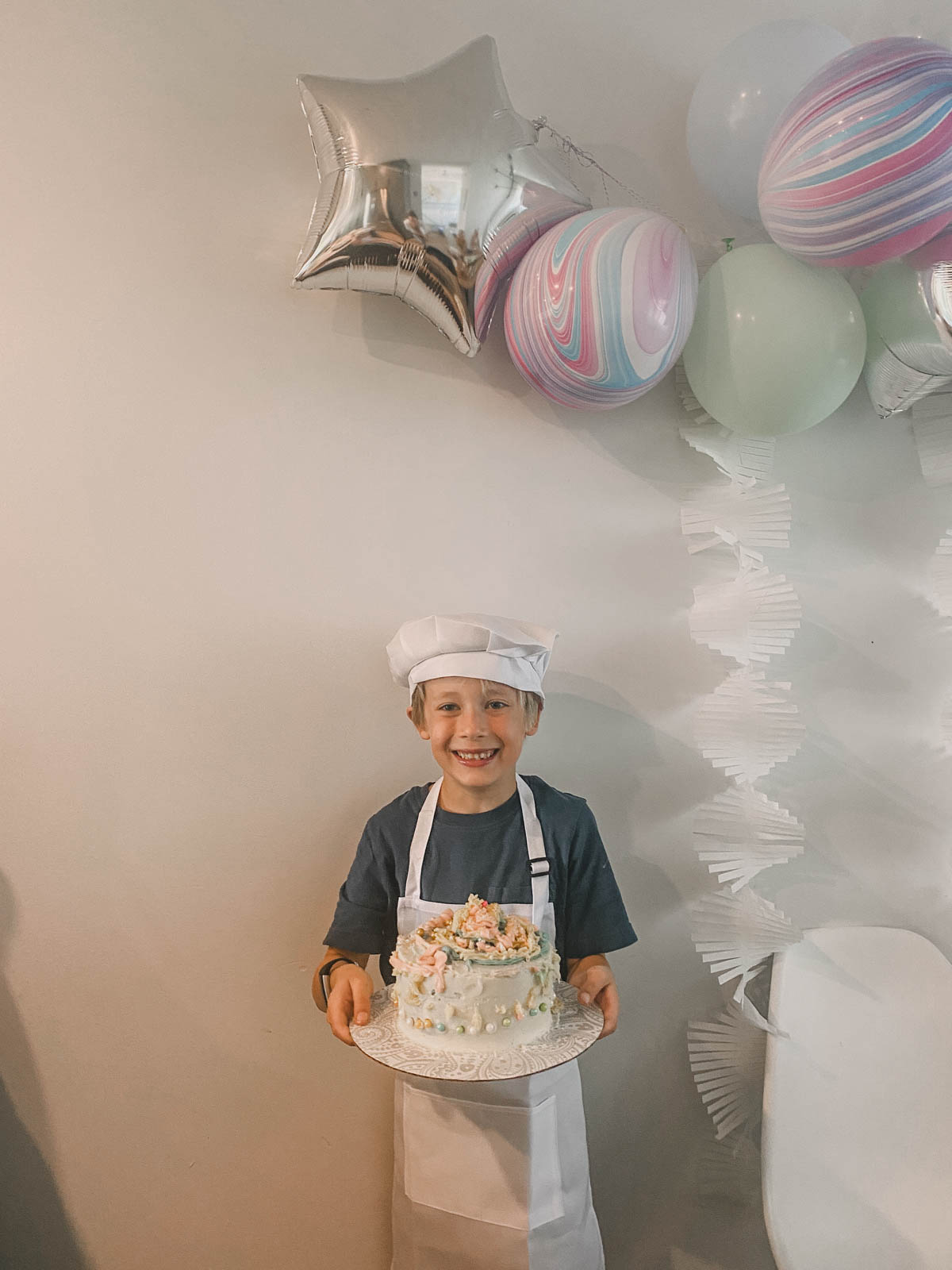 Boy holding a cake he decorated at a cake decorating party wearing an apron and chefs hat