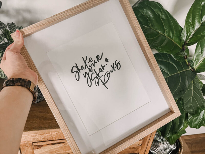 shake your shamrocks hand lettered art in a frame, held in front of plants
