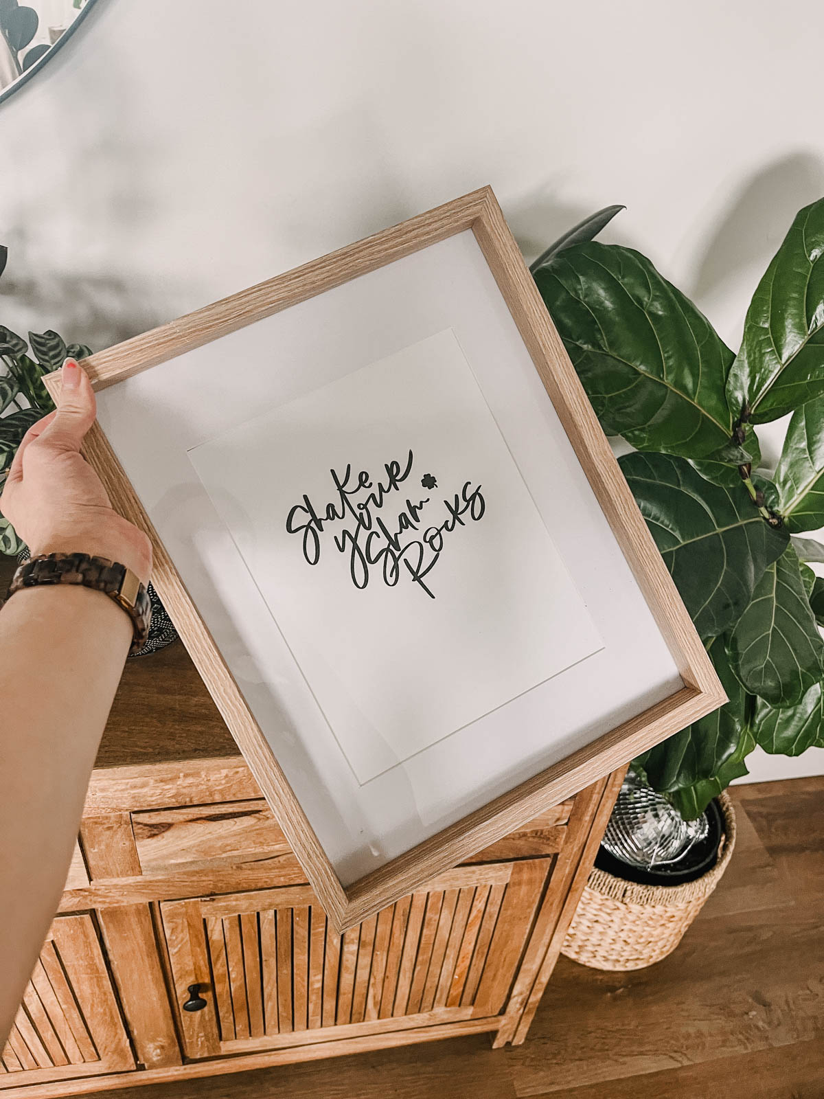 shake your shamrocks hand lettered art in a frame, held in front of plants