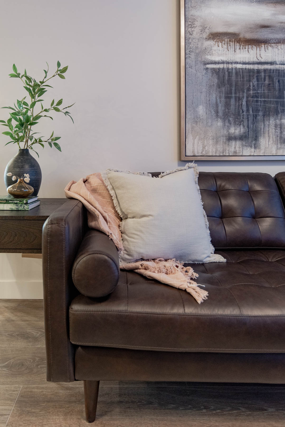 Simple spring decor- linen throw pillow and woven pink blanket on dark leather couch. Side table has small vase of dried bunny tail grasses and large vase of faux greenery