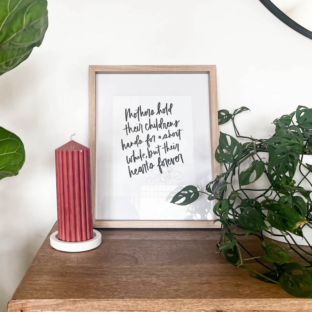 Hand lettered printable quote framed up for mothers day decor. Quote reads: mothers hold their childrens hands for a short while and their hearts forever