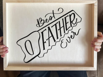 DIY wood sign being held by kid. Sign reads best father ever in saw outline, black vinyl on light wood panel.