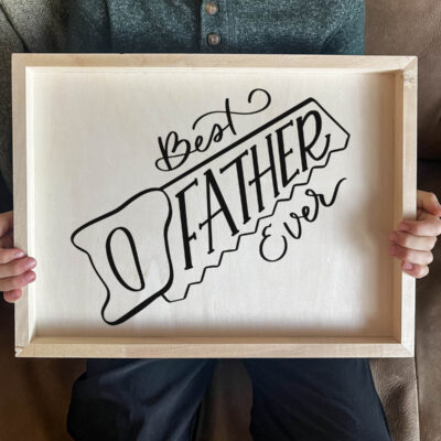 DIY wood sign being held by kid. Sign reads best father ever in saw outline, black vinyl on light wood panel.