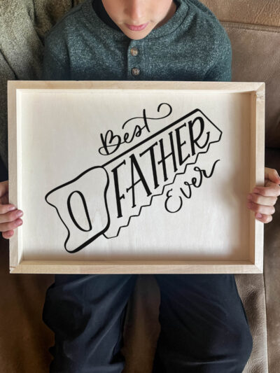 DIY wood sign being held by kid. Sign reads best father ever in saw outline, black vinyl on light wood panel.