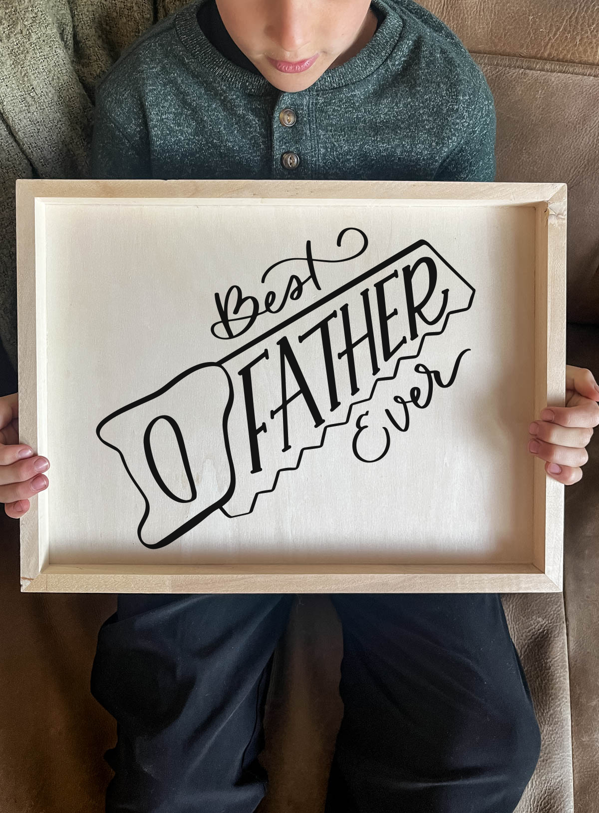 DIY wood sign being held by kid. Sign reads best father ever in saw outline, black vinyl on light wood panel. 
