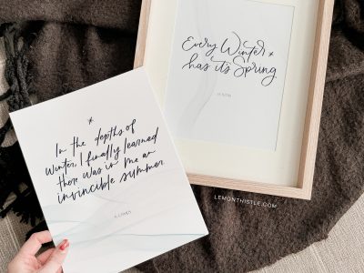 Free printable winter quotes shown printed on cardstock, one in wooden frame on cozy blanket