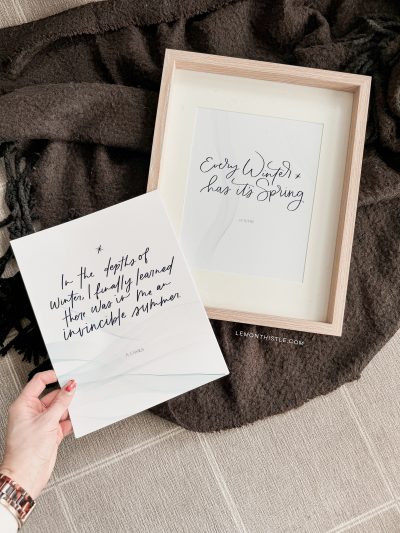 Free printable winter quotes shown printed on cardstock, one in wooden frame on cozy blanket