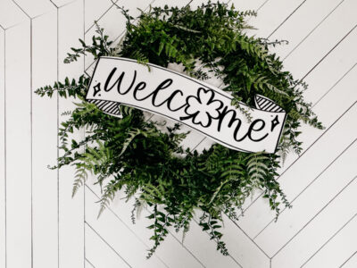 Image of fern leaf hung on geometric white barn door. Wreath features a large printed and cut to size welcome sign for saint patrick's day (free printable) welcome banner is hand drawn with brush marker style and shamrock O