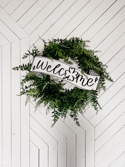 Image of fern leaf hung on geometric white barn door. Wreath features a large printed and cut to size welcome sign for saint patrick's day (free printable) welcome banner is hand drawn with brush marker style and shamrock O