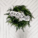 Image of fern leaf hung on geometric white barn door. Wreath features a large printed and cut to size welcome sign for saint patrick's day (free printable) welcome banner is hand drawn with brush marker style and shamrock O