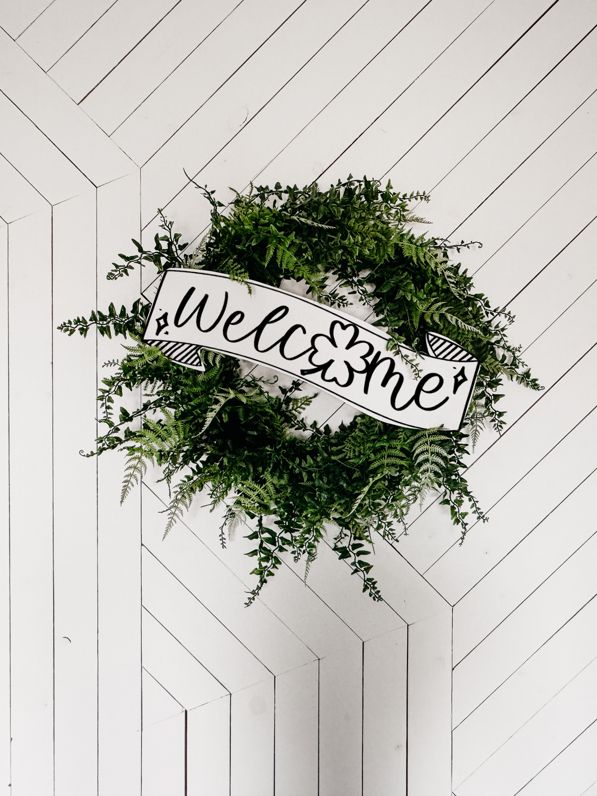 Image of fern leaf hung on geometric white barn door. Wreath features a large printed and cut to size welcome sign for saint patrick's day (free printable) welcome banner is hand drawn with brush marker style and shamrock O