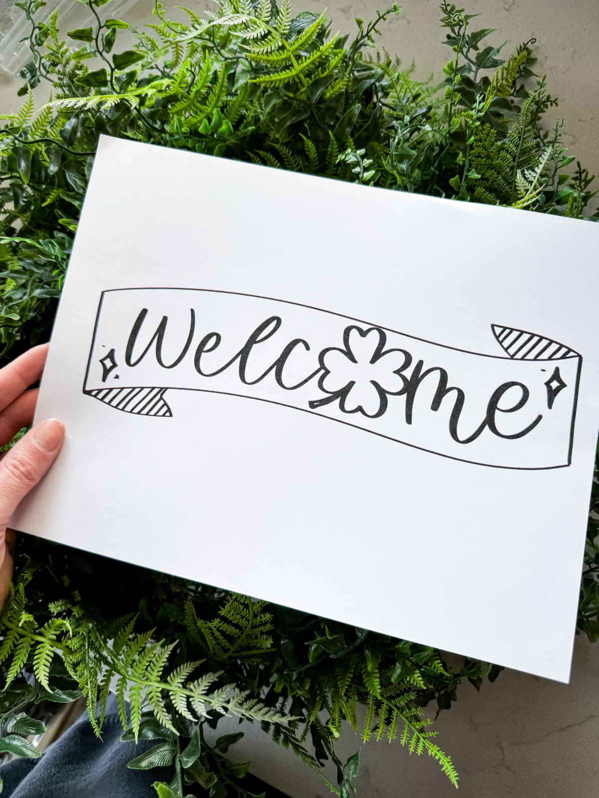 Image of hand holding a printed welcome banner on 8x5x11 paper banner is hand drawn with a brush marker style, printed in black and white. the banners ays welcome with the O as a shamrock printed banner is being held over a fern leaf