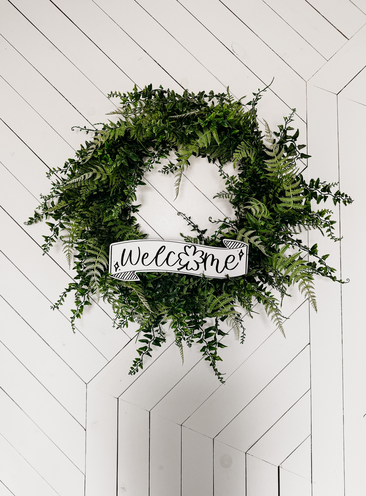 Image of fern leaf hung on geometric white barn door. Wreath features a printed and cut to size welcome sign for saint patrick's day (free printable) welcome banner is hand drawn with brush marker style and shamrock O
