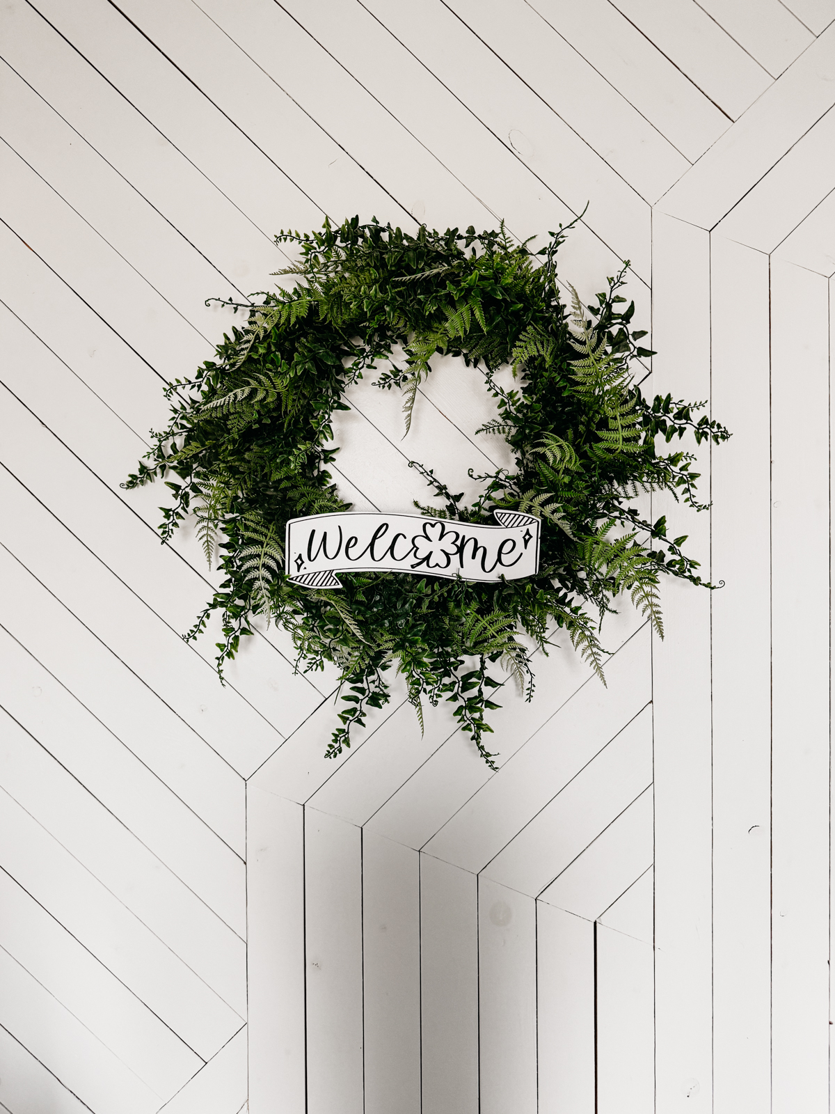 Image of fern leaf hung on geometric white barn door. Wreath features a printed and cut to size welcome sign for saint patrick's day (free printable) welcome banner is hand drawn with brush marker style and shamrock O
