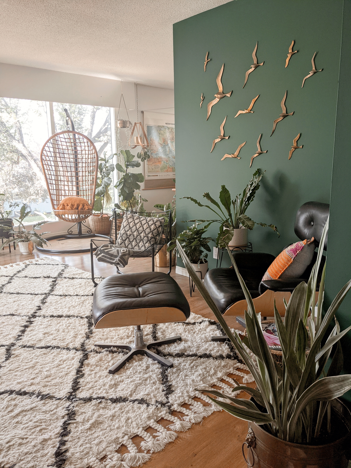 Living room full of potted plants, green accent wall features brass birds, rattan hanging chair in background