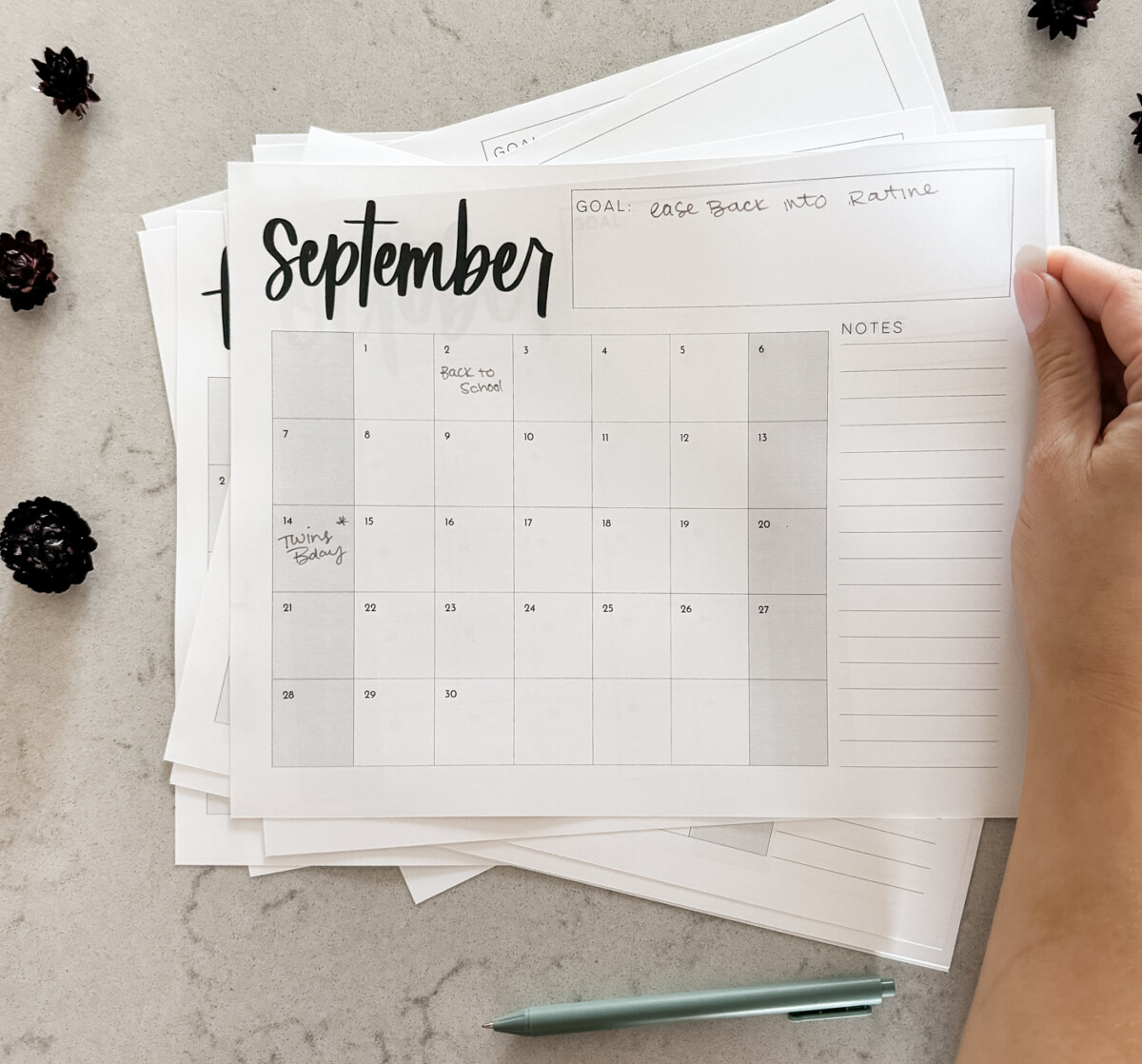monthly calendar with grid format and room for notes. calendar month features hand lettering and a minimalist style, shown printed on a marble countertop with green pen, being held by hand