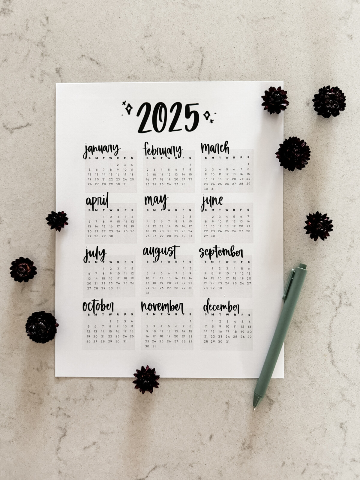 2025 year at a glance calendar shown printed on marble countertop with dark flowers. Hand lettered details and minimalist style
